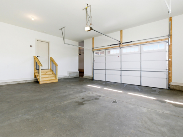 Bright, empty residential garage with white walls, a small wooden staircase to a door, and a large metal sectional garage door on the right.