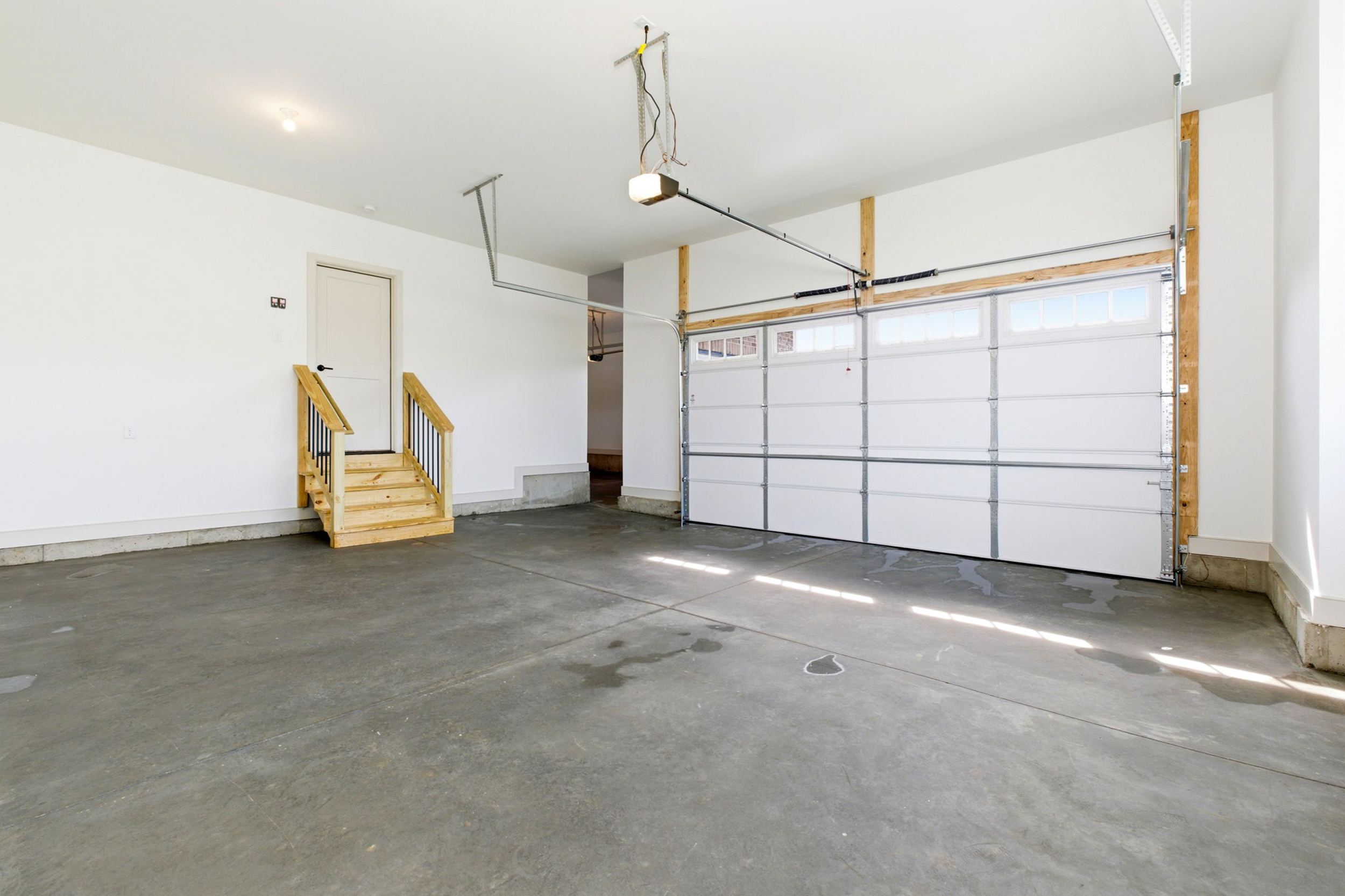 Bright, empty residential garage with white walls, a small wooden staircase to a door, and a large metal sectional garage door on the right.