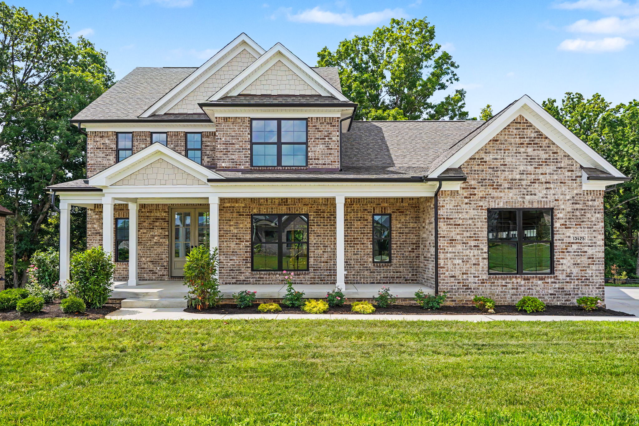 13425 Riggs Lake Ln-01 Brick two-story house with a covered front porch, large windows, and a manicured lawn under a blue sky with green trees in the background.