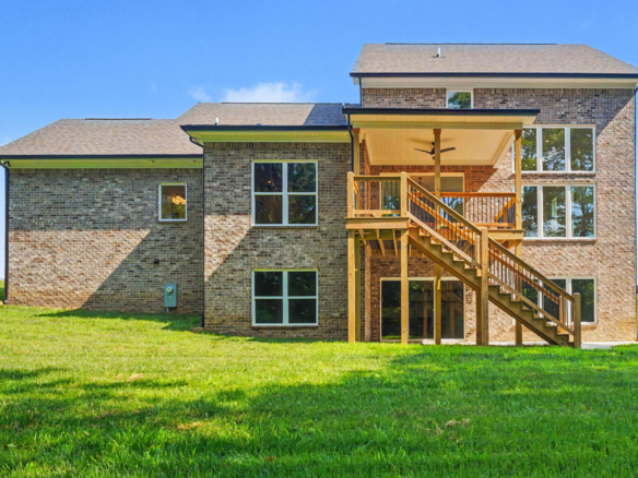 Rear view of a brick two-story house with a large wooden deck and external stairs, green lawn and a clear blue sky.