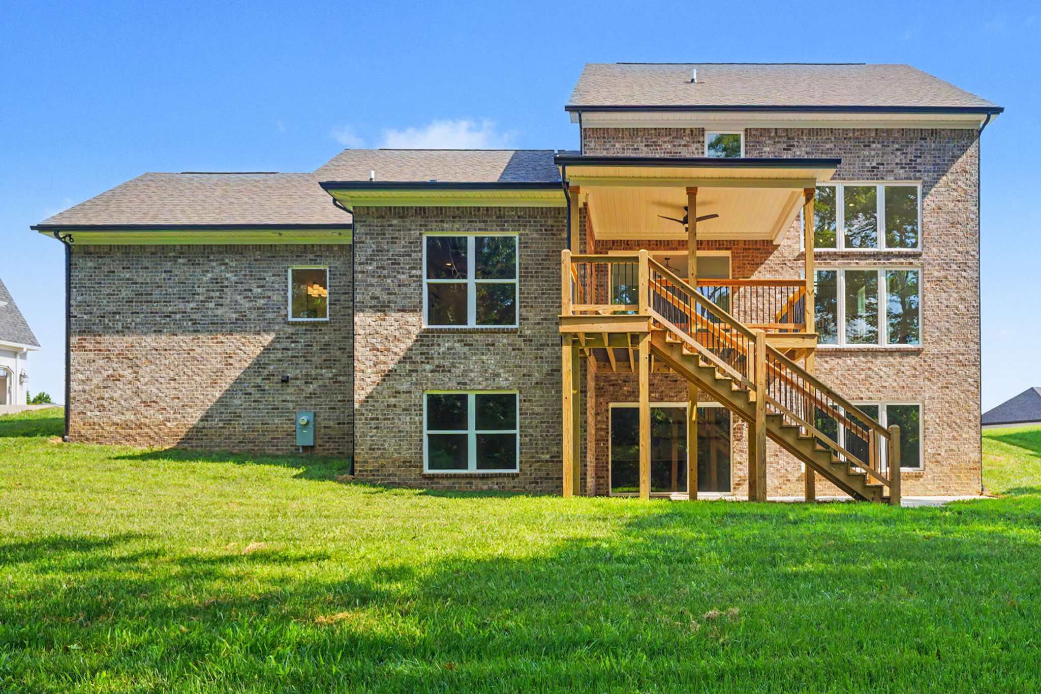Rear view of a brick two-story house with a large wooden deck and external stairs, green lawn and a clear blue sky.