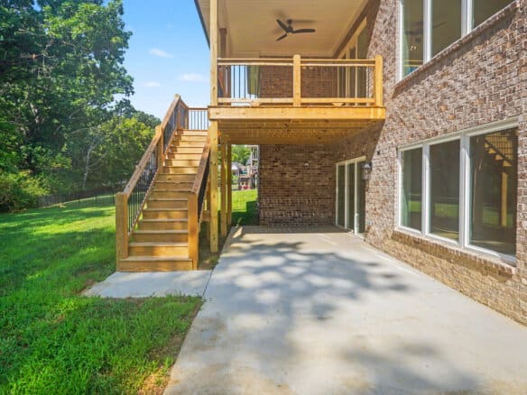 Backyard view of a brick house with a wooden deck and stairs, concrete patio, and green lawn.