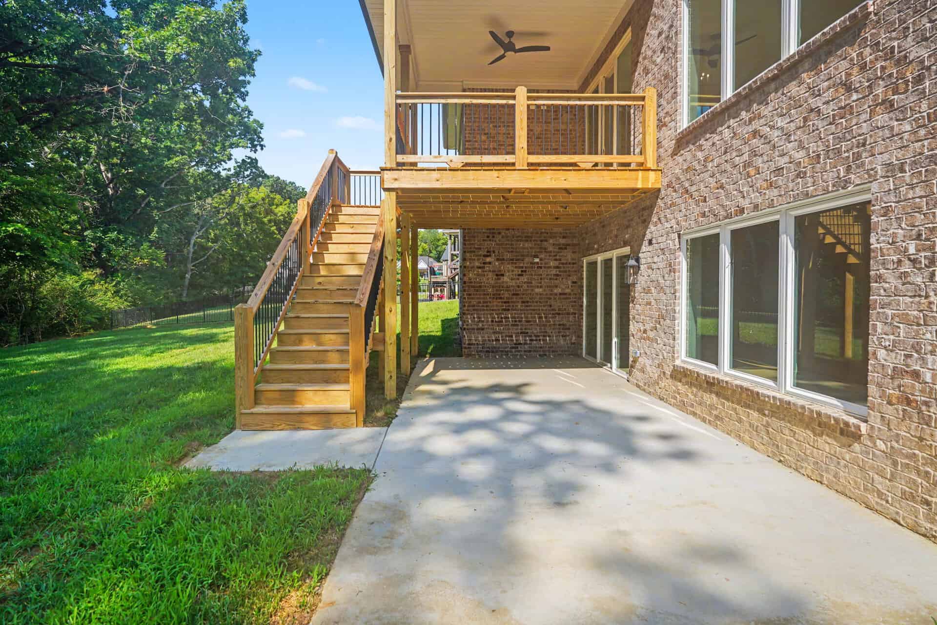 Backyard view of a brick house with a wooden deck and stairs, concrete patio, and green lawn.