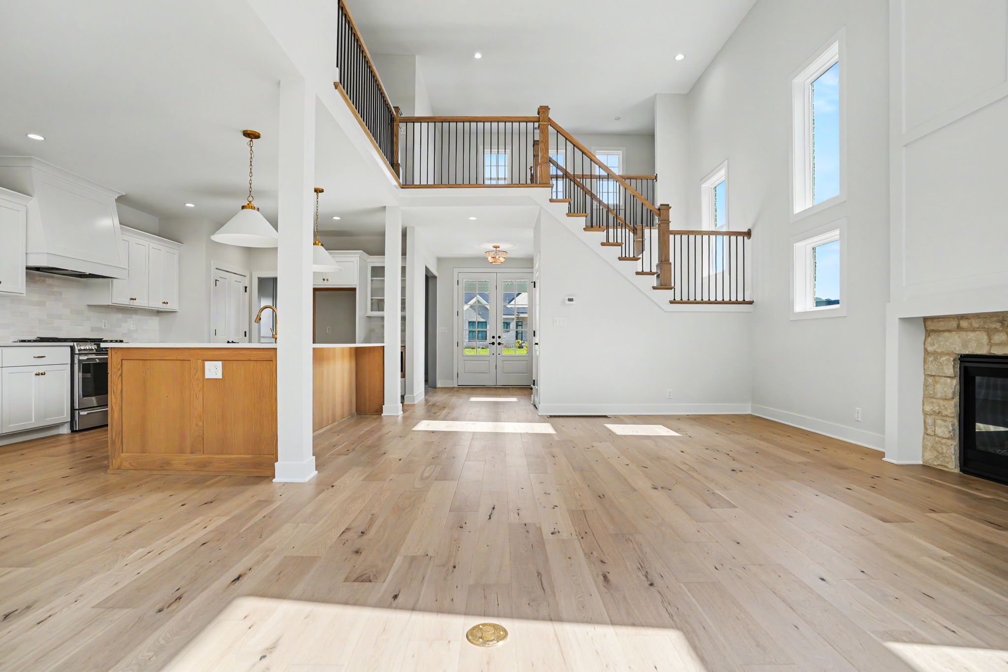 Bright open-concept living space featuring a white kitchen to the left, a central island, and a wooden staircase with black balusters leading to the second floor.