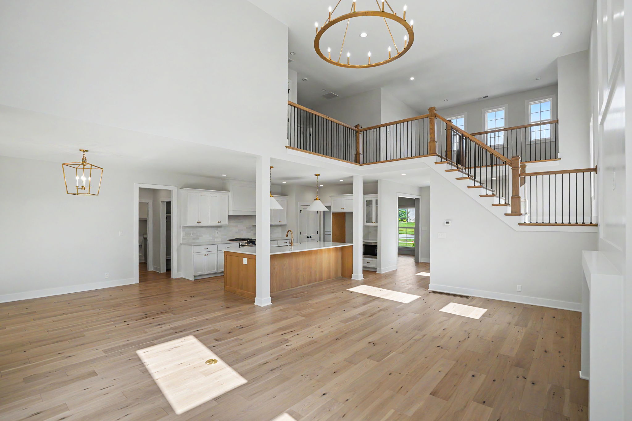 Bright, two-story open-plan living area with white walls, a wooden staircase with black balusters, and a gold circular chandelier.