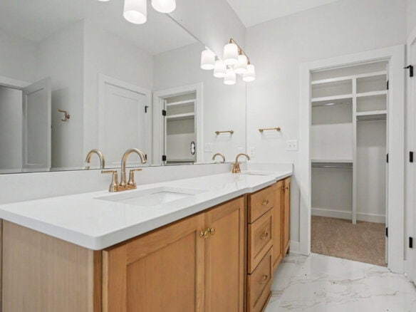 Double-sink bathroom vanity with light gray countertop, brass fixtures, and wooden cabinetry under a large mirror in a bright, modern bathroom.