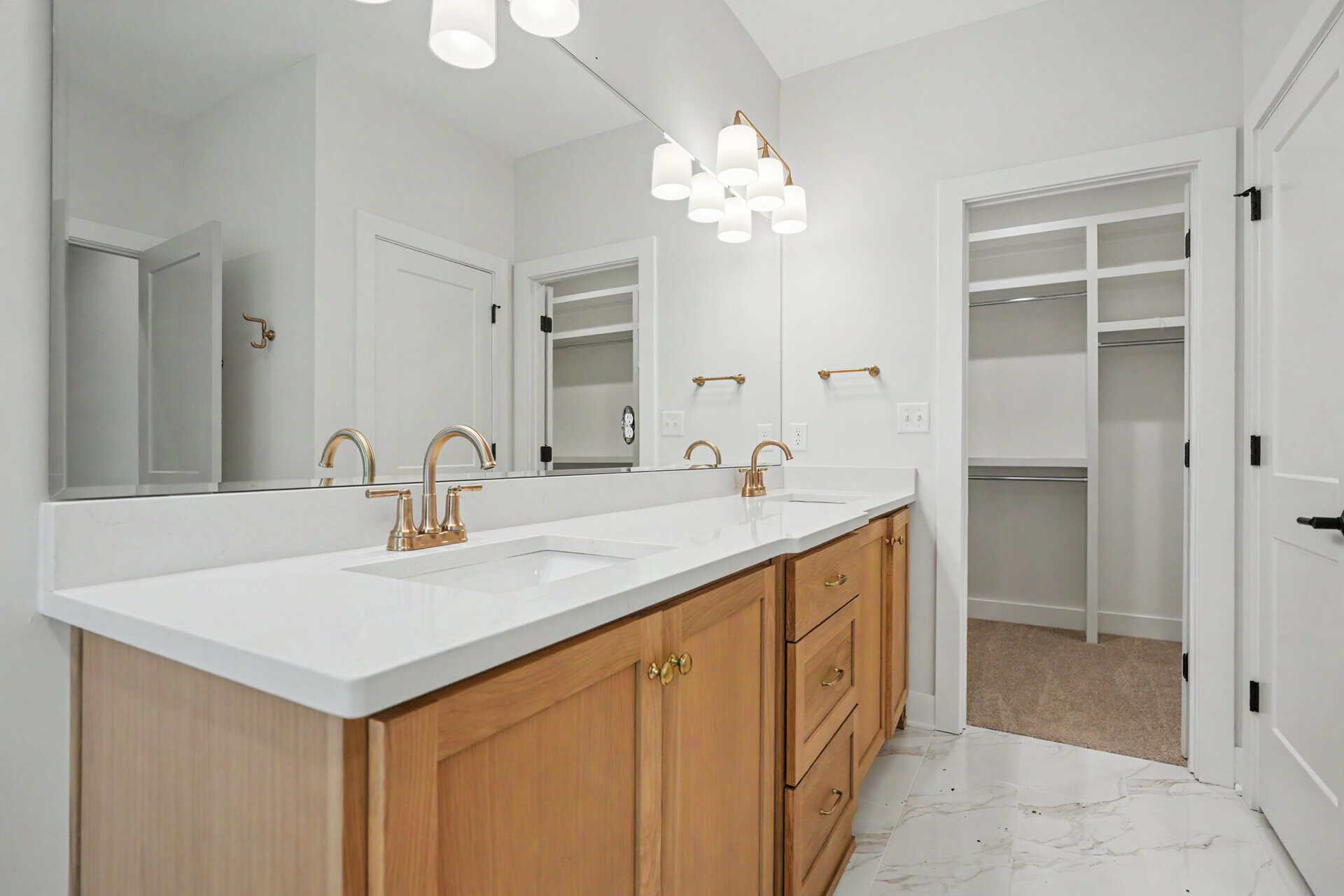 Double-sink bathroom vanity with light gray countertop, brass fixtures, and wooden cabinetry under a large mirror in a bright, modern bathroom.