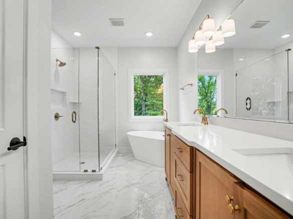 Bright modern bathroom with a double vanity, wooden cabinets, and brass fixtures beside a glass-enclosed shower.