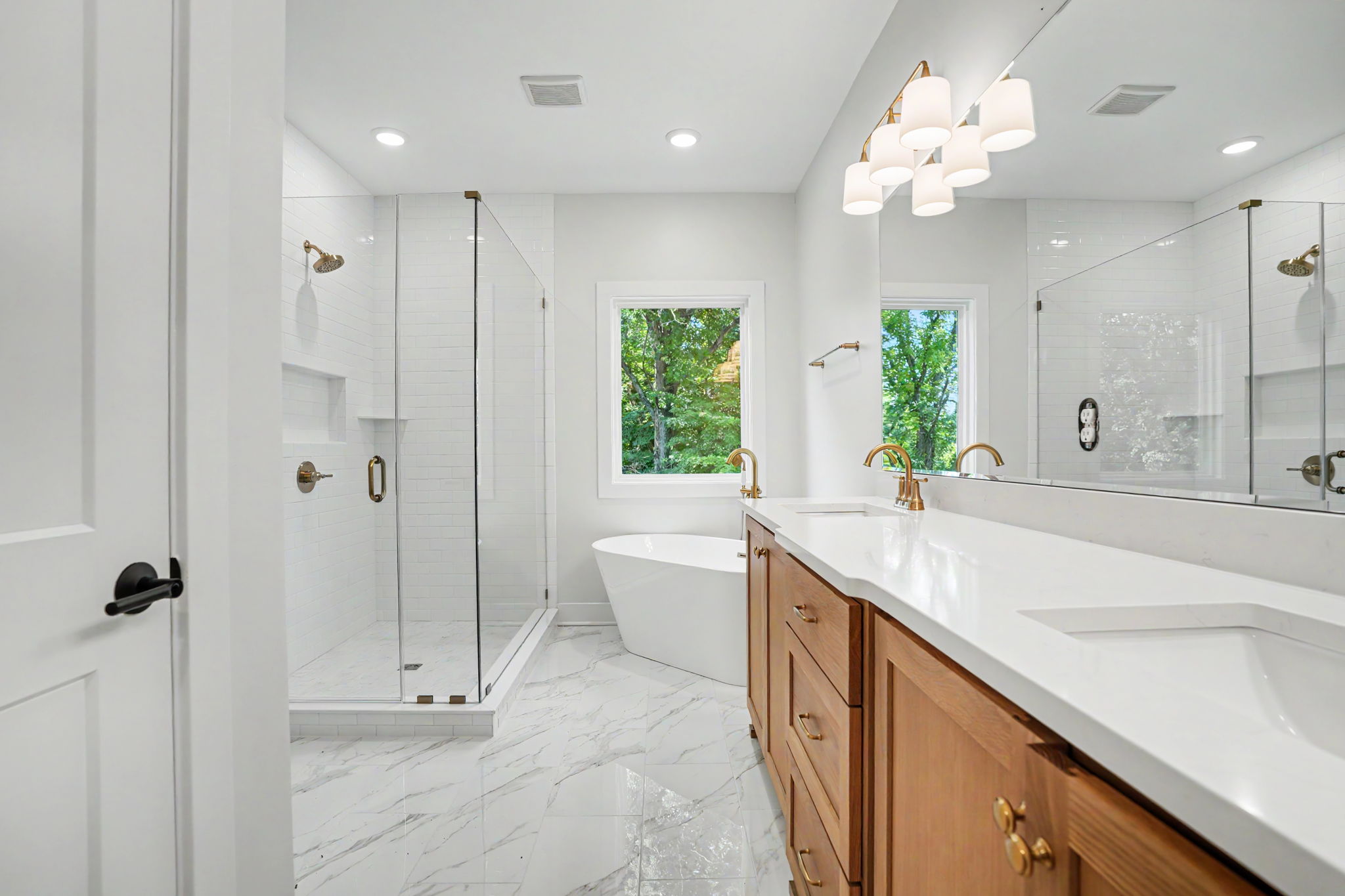 Bright modern bathroom with a double vanity, wooden cabinets, and brass fixtures beside a glass-enclosed shower.