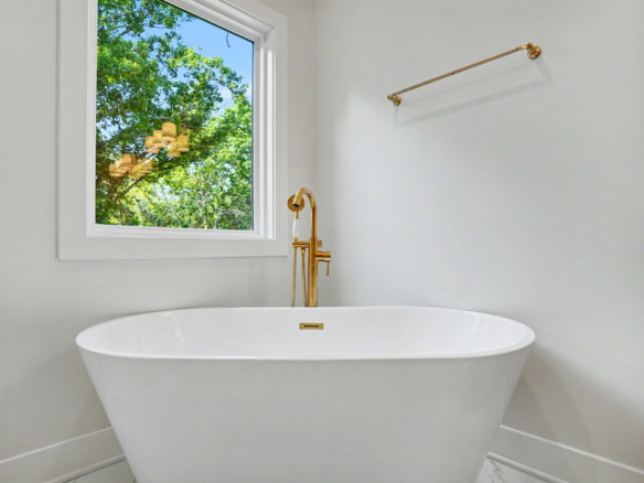 White freestanding bathtub with a gold faucet in a bright bathroom, window showing green trees outside