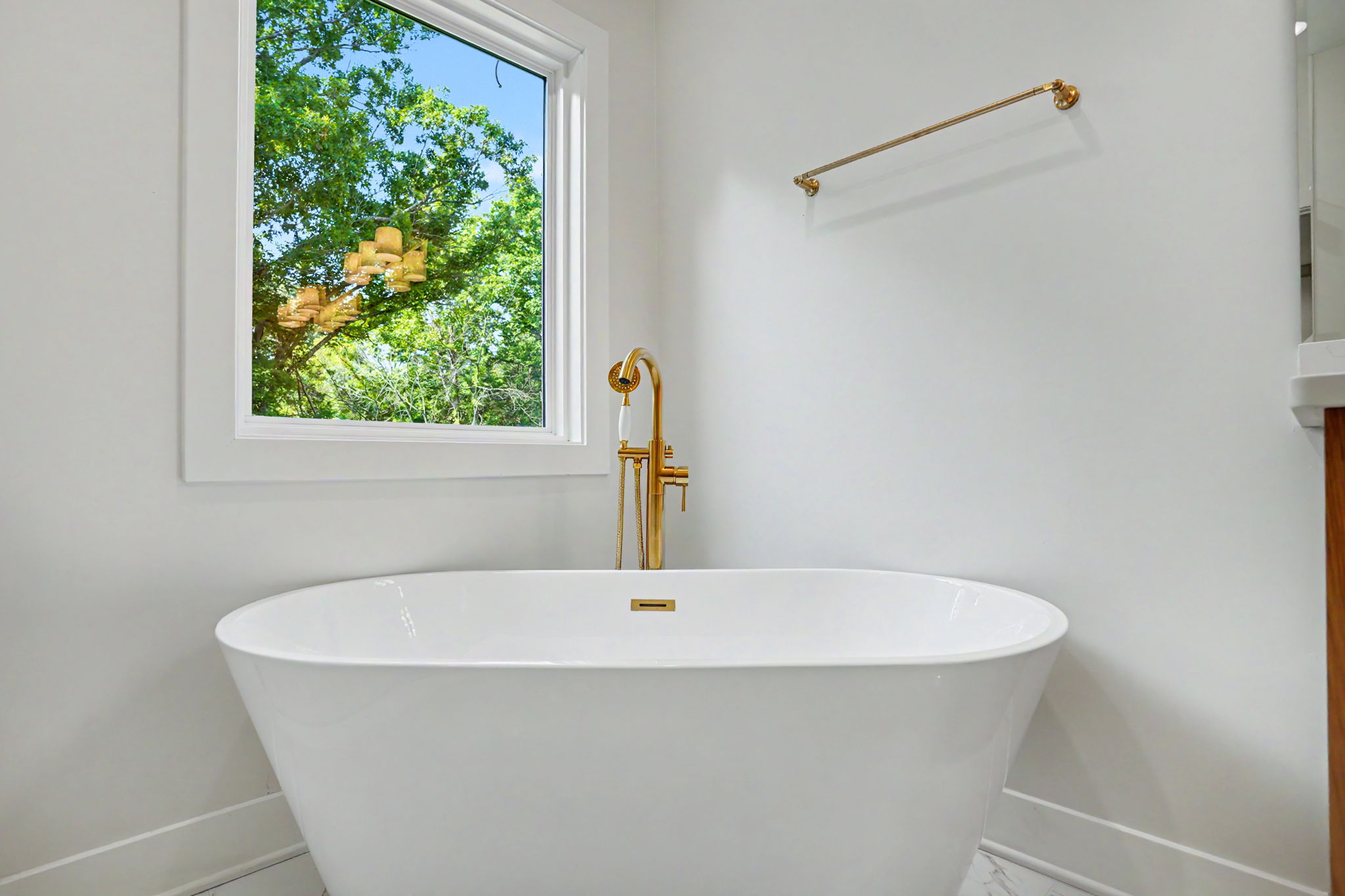 White freestanding bathtub with a gold faucet in a bright bathroom, window showing green trees outside