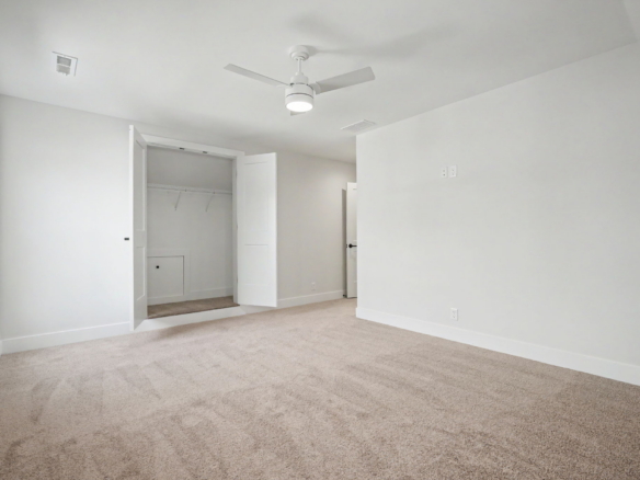 Empty bedroom with beige carpet, white walls, open closet doors, and a white ceiling fan in the center ceiling.