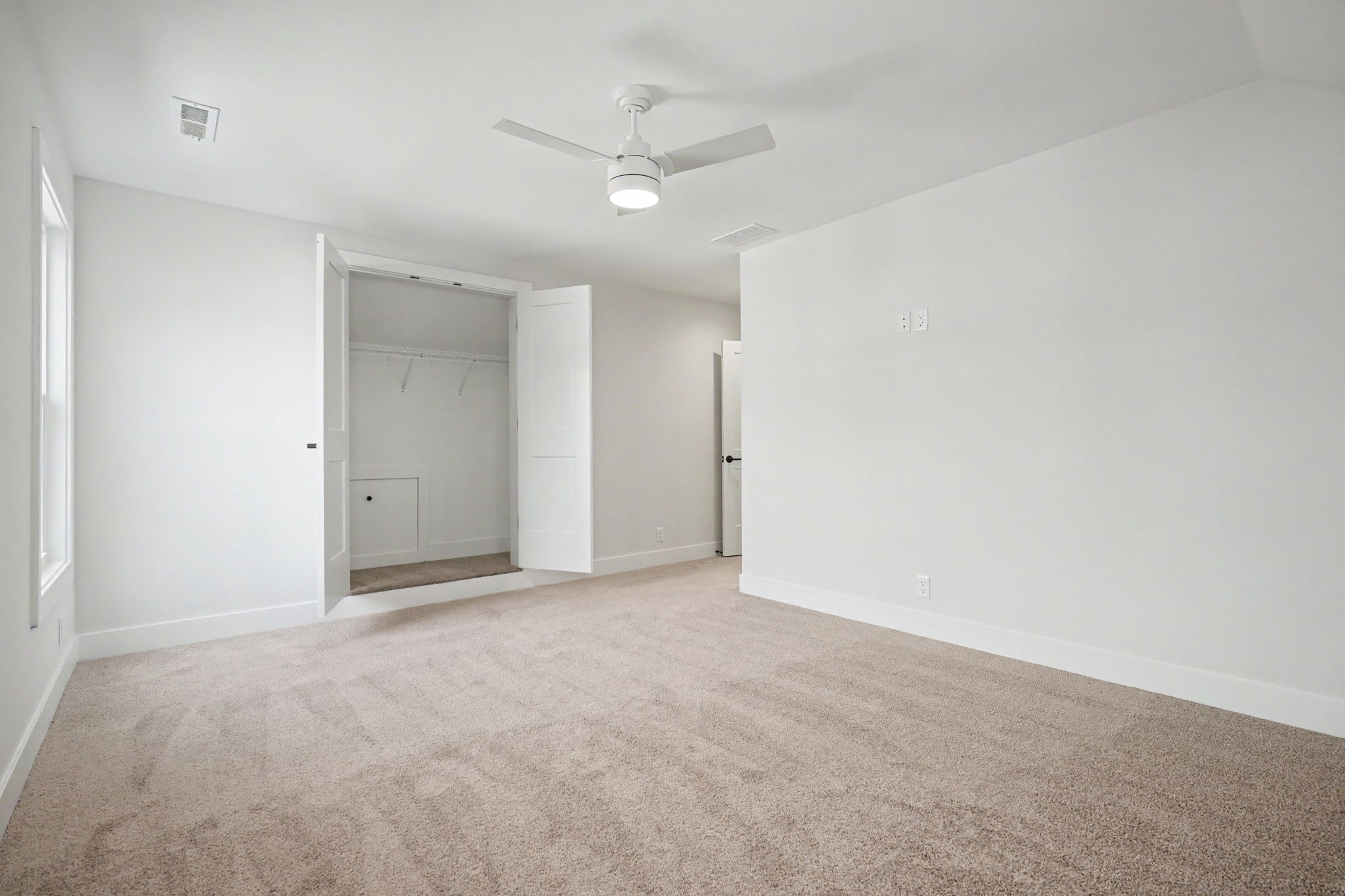 Empty bedroom with beige carpet, white walls, open closet doors, and a white ceiling fan in the center ceiling.