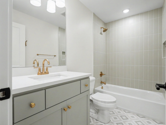 Modern bathroom with gray vanity, white countertop, and gold faucet; toilet and tiled shower area with brass fixtures nearby.
