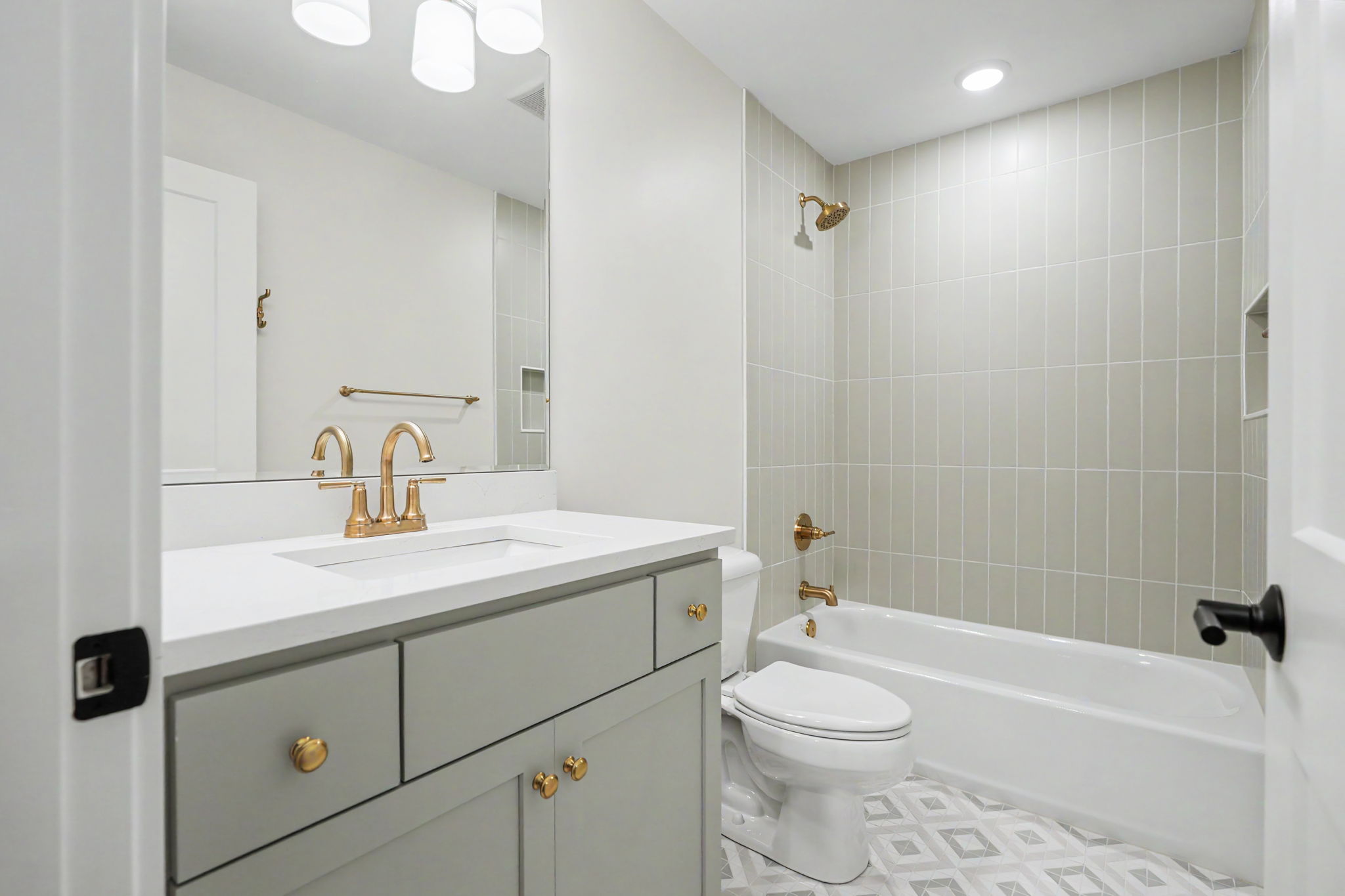 Modern bathroom with gray vanity, white countertop, and gold faucet; toilet and tiled shower area with brass fixtures nearby.