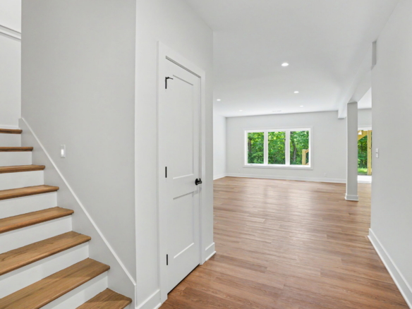 Bright modern entry hall with wooden stairs on the left, white walls, and a doorway leading to a living space with a large window showing green trees outside.