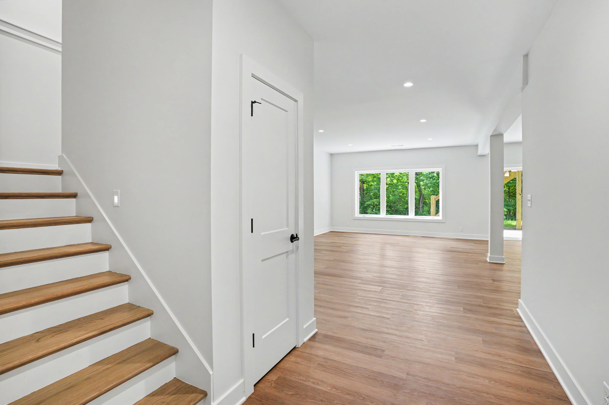 Bright modern entry hall with wooden stairs on the left, white walls, and a doorway leading to a living space with a large window showing green trees outside.