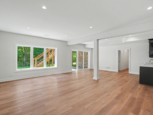 Bright open living space with light wood flooring, large windows showing greenery, and an adjacent kitchen island on the right.