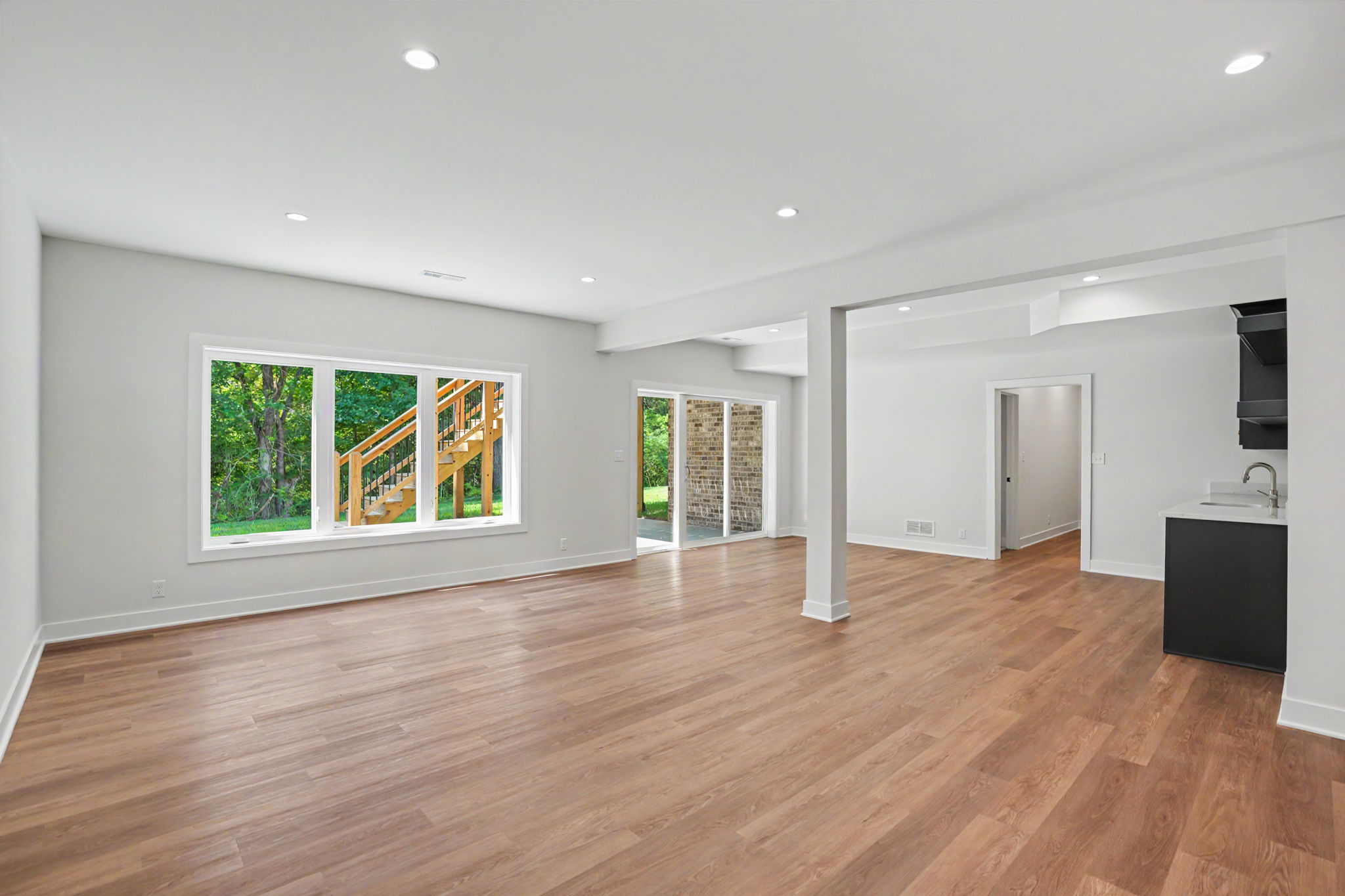 Bright open living space with light wood flooring, large windows showing greenery, and an adjacent kitchen island on the right.
