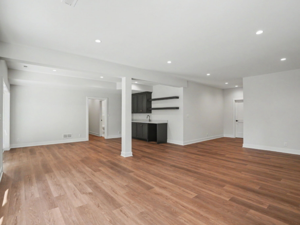 Spacious open-concept living area with white walls, hardwood floors, and recessed ceiling lights; a dark-kitchen island with cabinets and open shelves is visible in the background.