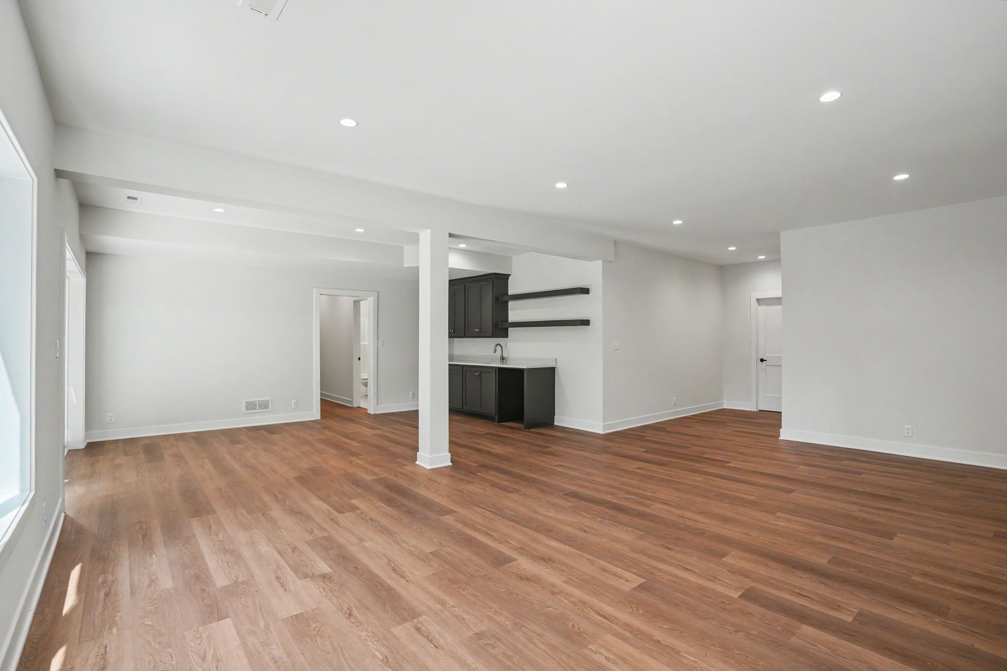 Spacious open-concept living area with white walls, hardwood floors, and recessed ceiling lights; a dark-kitchen island with cabinets and open shelves is visible in the background.