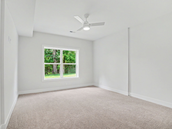 Bright empty room with light gray walls, beige carpet, and a white ceiling fan; window shows green trees outside