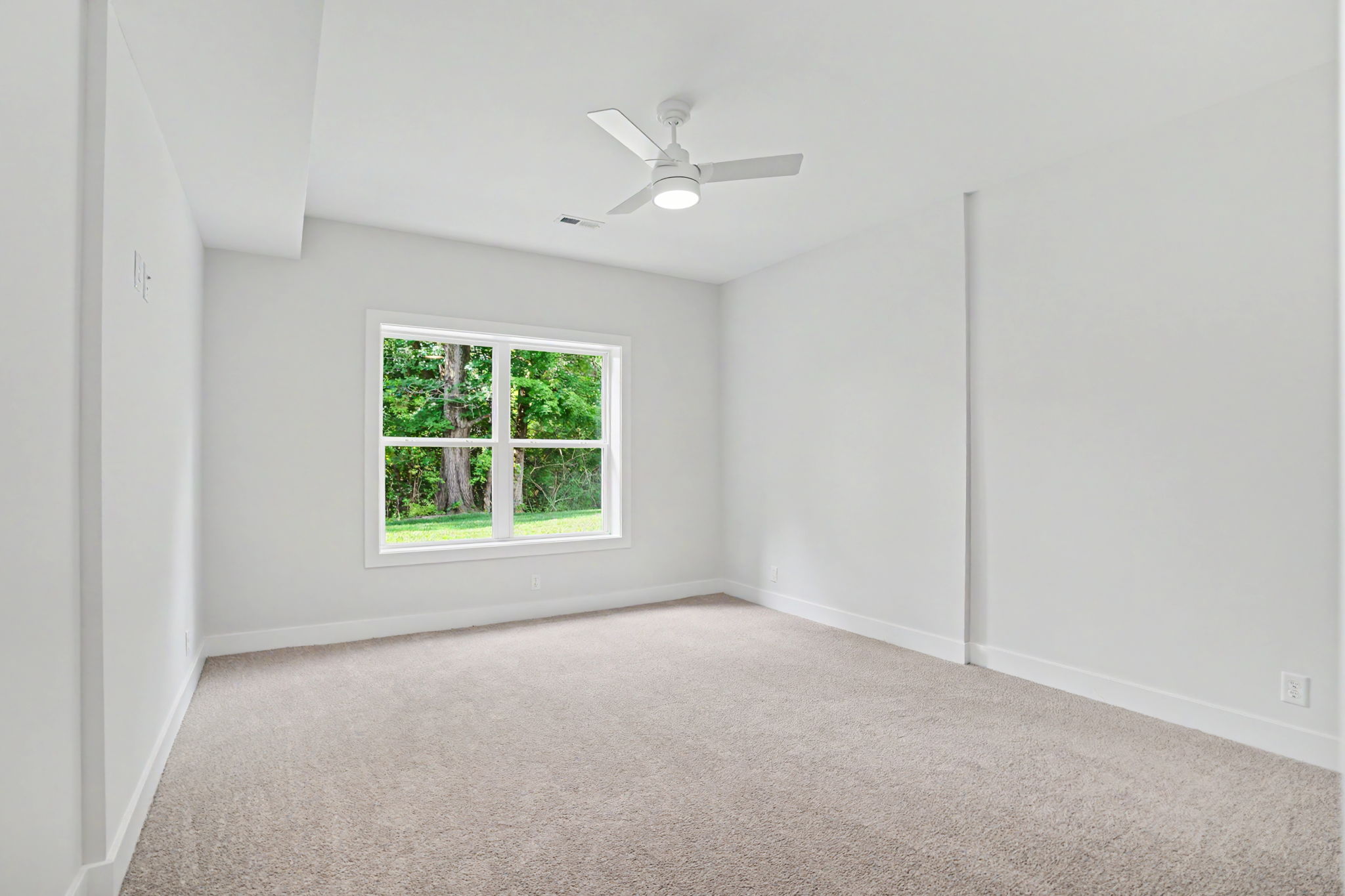 Bright empty room with light gray walls, beige carpet, and a white ceiling fan; window shows green trees outside