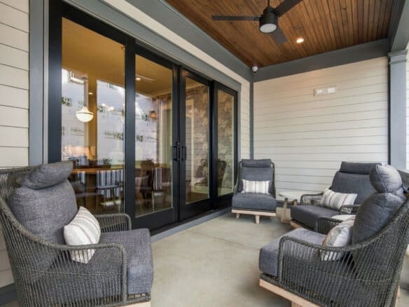 Covered outdoor patio with gray wicker chairs and striped cushions facing glass sliding doors. Ceiling fan above.