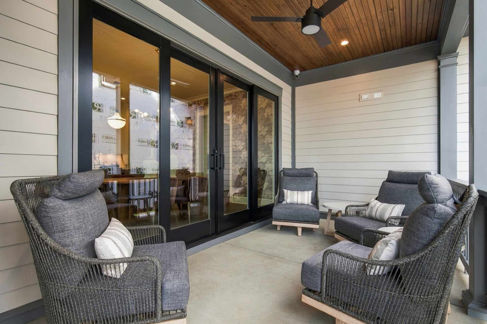 Covered outdoor patio with gray wicker chairs and striped cushions facing glass sliding doors. Ceiling fan above.