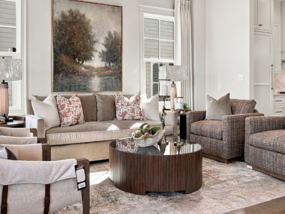 Bright, neutral-toned living room with beige sofa, patterned throw pillows, and a round ribbed wood coffee table; large landscape painting on the wall and windows with curtains in the background.