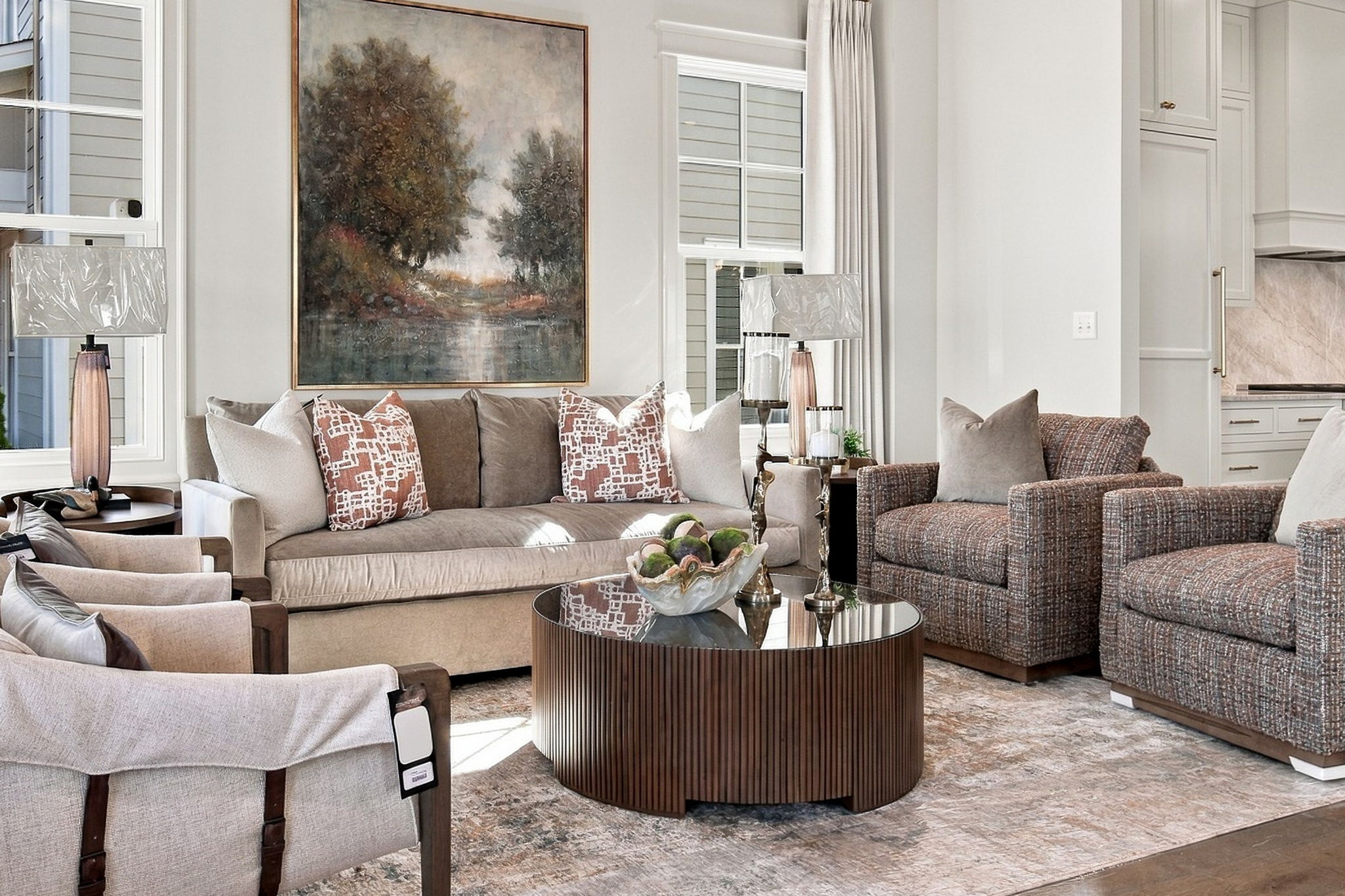 Bright, neutral-toned living room with beige sofa, patterned throw pillows, and a round ribbed wood coffee table; large landscape painting on the wall and windows with curtains in the background.