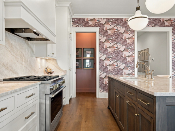 Bright kitchen with white cabinetry, marble countertops, and a dark wood island beside a pink floral wallpapered wall.