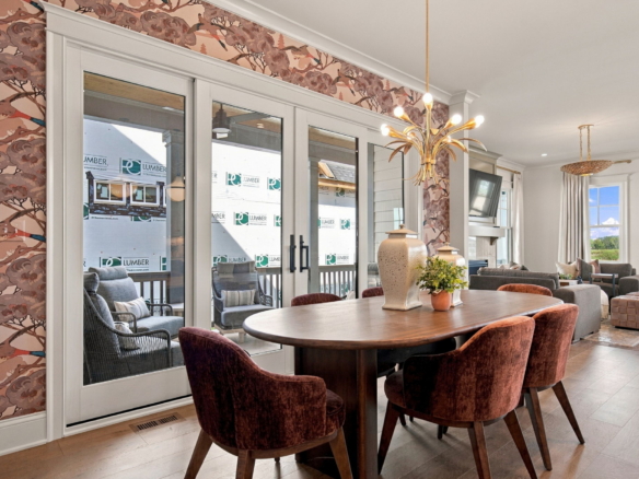 Round wooden dining table with burgundy upholstered chairs in an open living/dining space, pink patterned wallpaper with birds, and a gold palm-shaped chandelier hanging overhead; sliding glass doors to an outdoor area.