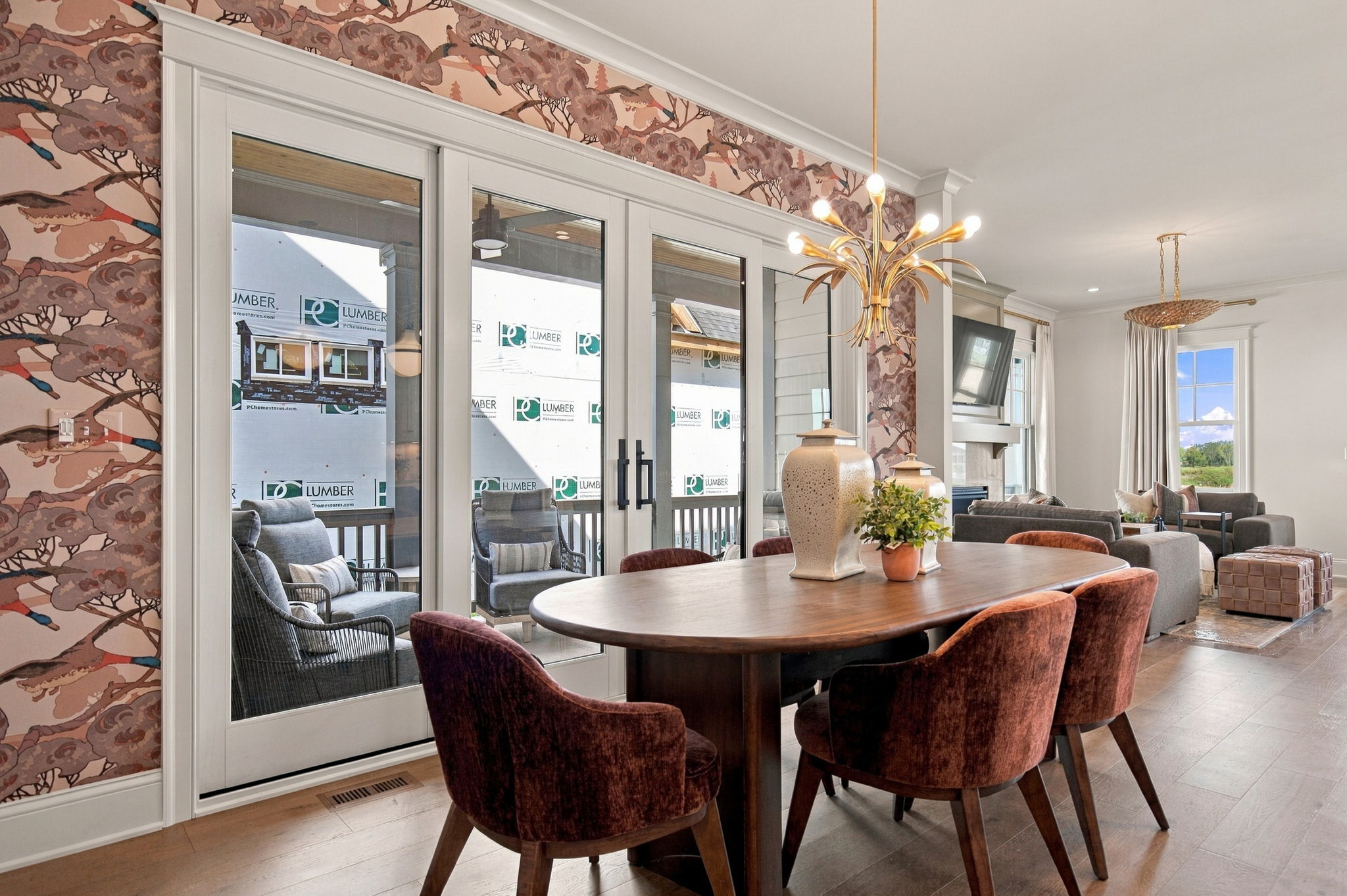 Round wooden dining table with burgundy upholstered chairs in an open living/dining space, pink patterned wallpaper with birds, and a gold palm-shaped chandelier hanging overhead; sliding glass doors to an outdoor area.