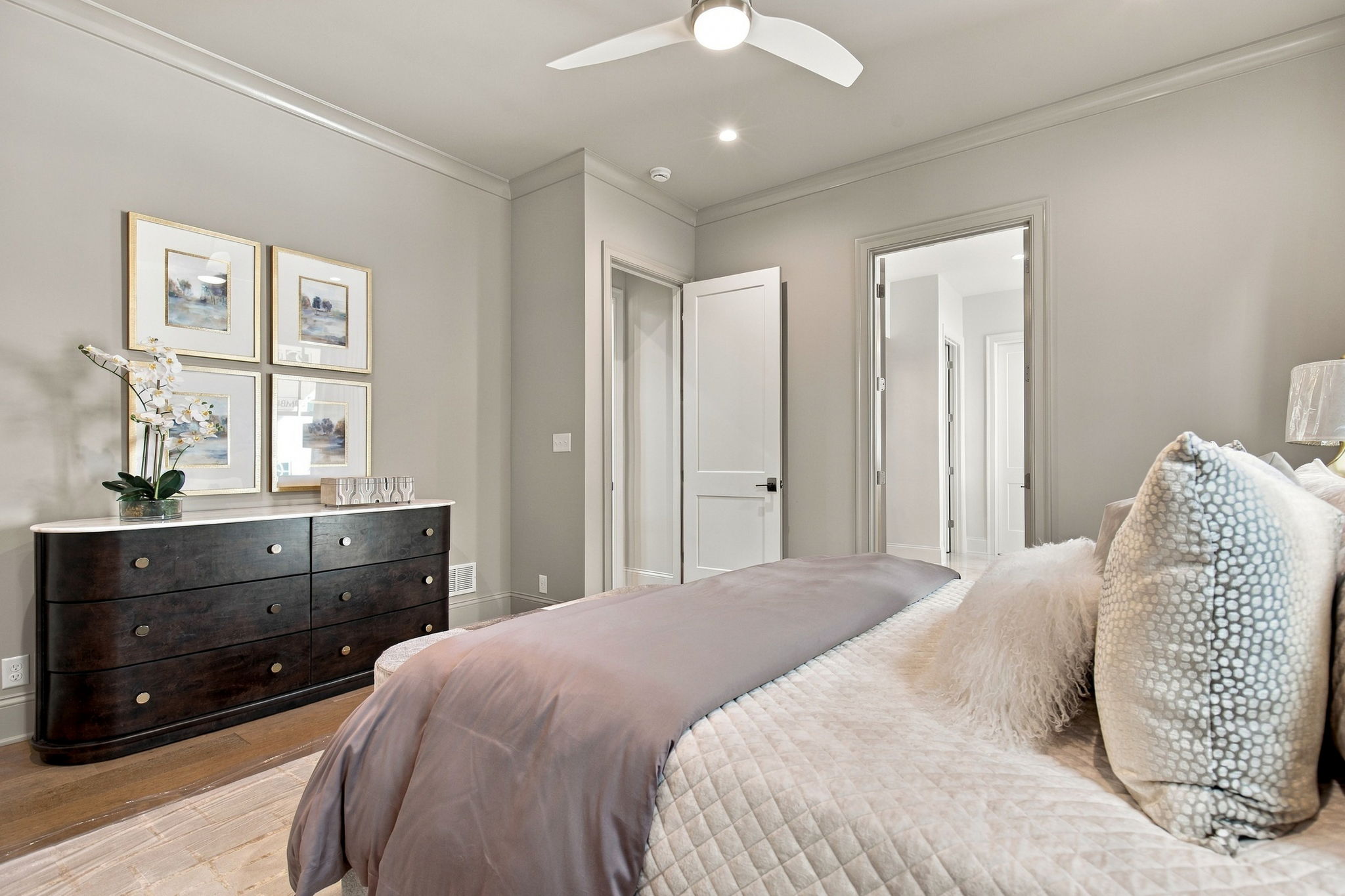 Bedroom with taupe bedding, a dark wood dresser, and four framed gold-framed artworks on a light gray wall.