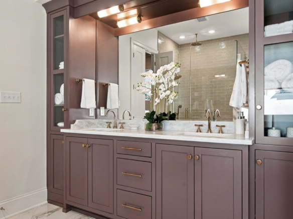 Purple bathroom vanity with marble countertop, gold faucets, and a large mirror; white towels hang on each side and an orchid centerpiece on the counter.