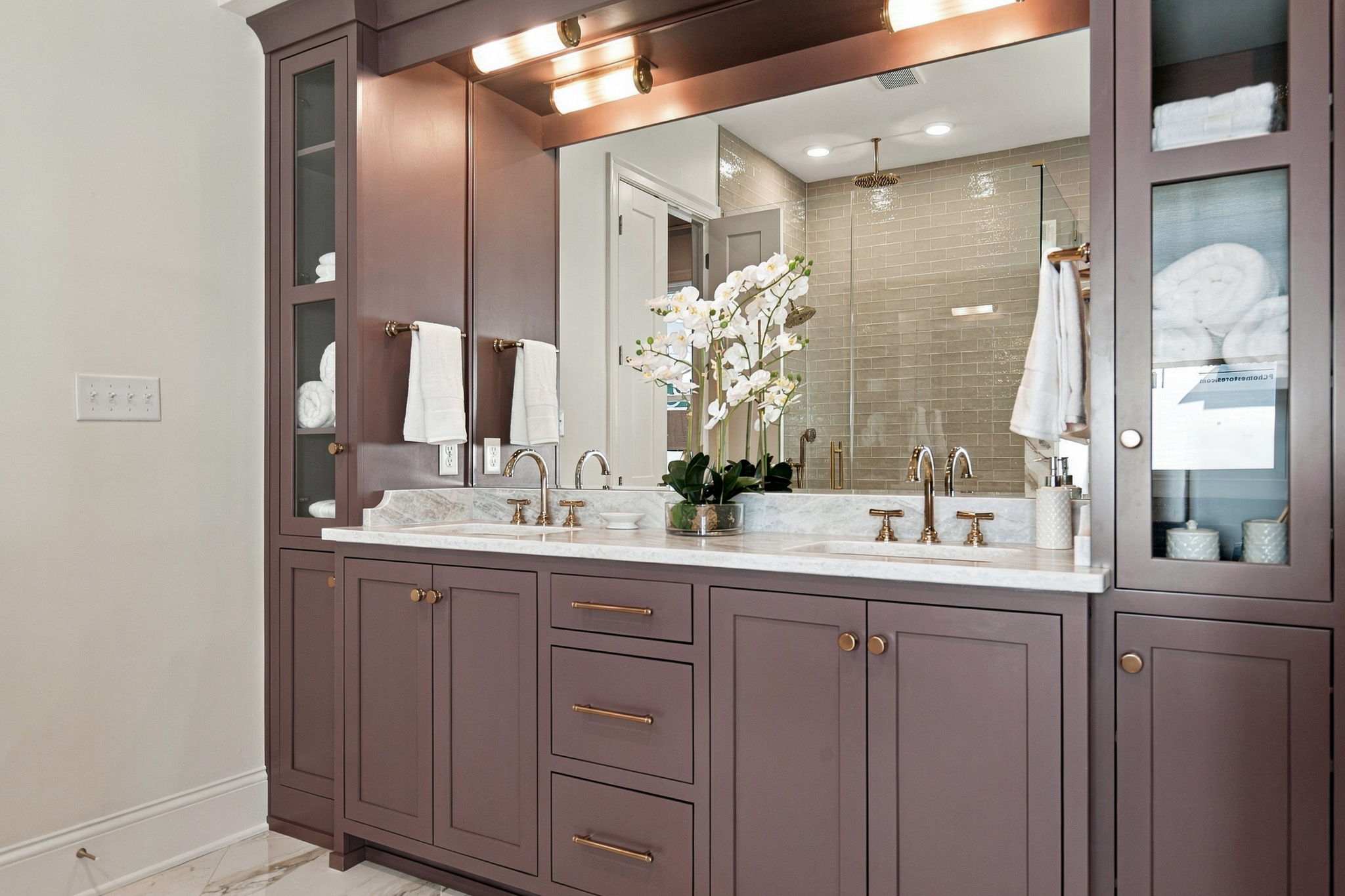 Purple bathroom vanity with marble countertop, gold faucets, and a large mirror; white towels hang on each side and an orchid centerpiece on the counter.