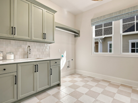 Bright kitchen with sage-grey cabinets, white tile backsplash, and a stainless sink under a large window with a fabric valance, plus a checkered tile floor.