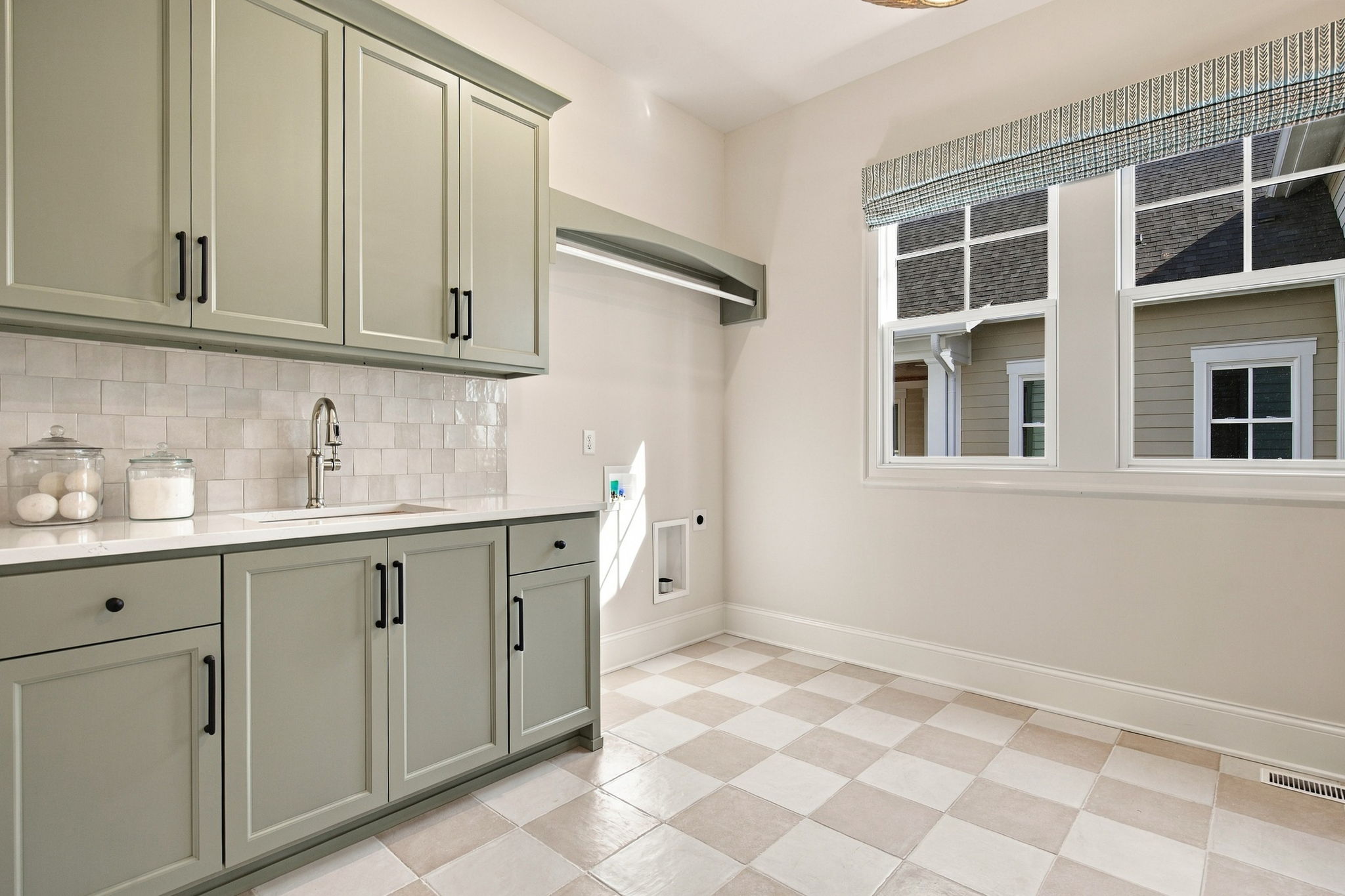 Bright kitchen with sage-grey cabinets, white tile backsplash, and a stainless sink under a large window with a fabric valance, plus a checkered tile floor.