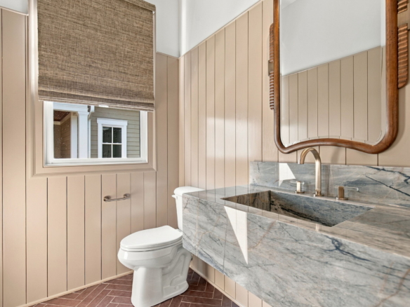 Bathroom featuring beige beadboard walls, marble countertop, curved wooden-framed mirror, window with shade, and a toilet.