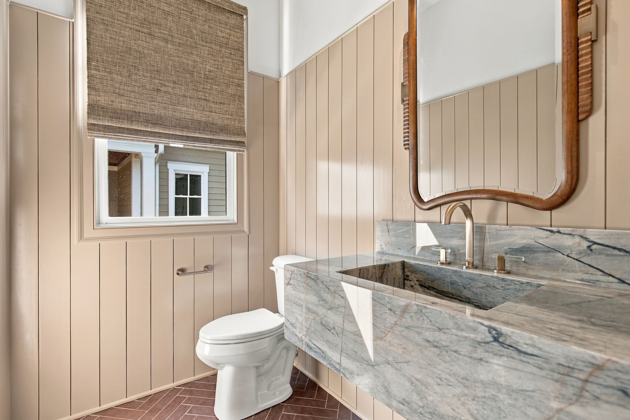 Bathroom featuring beige beadboard walls, marble countertop, curved wooden-framed mirror, window with shade, and a toilet.