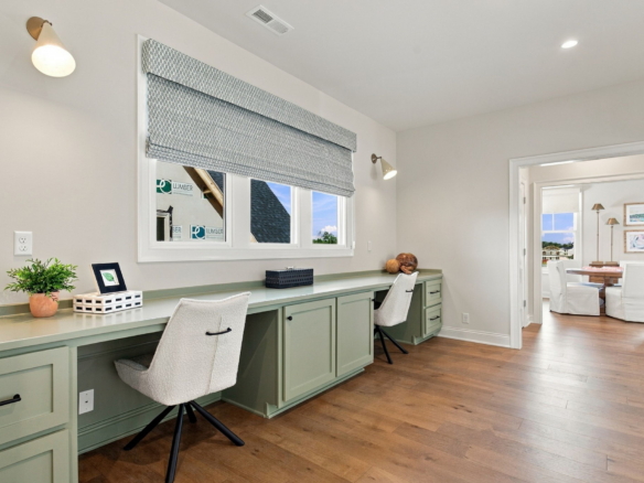 Long mint-green built-in desk with matching cabinets runs along a sunlit wall, two white swivel chairs, a potted plant, and decorative items under a patterned window shade in a bright home office.