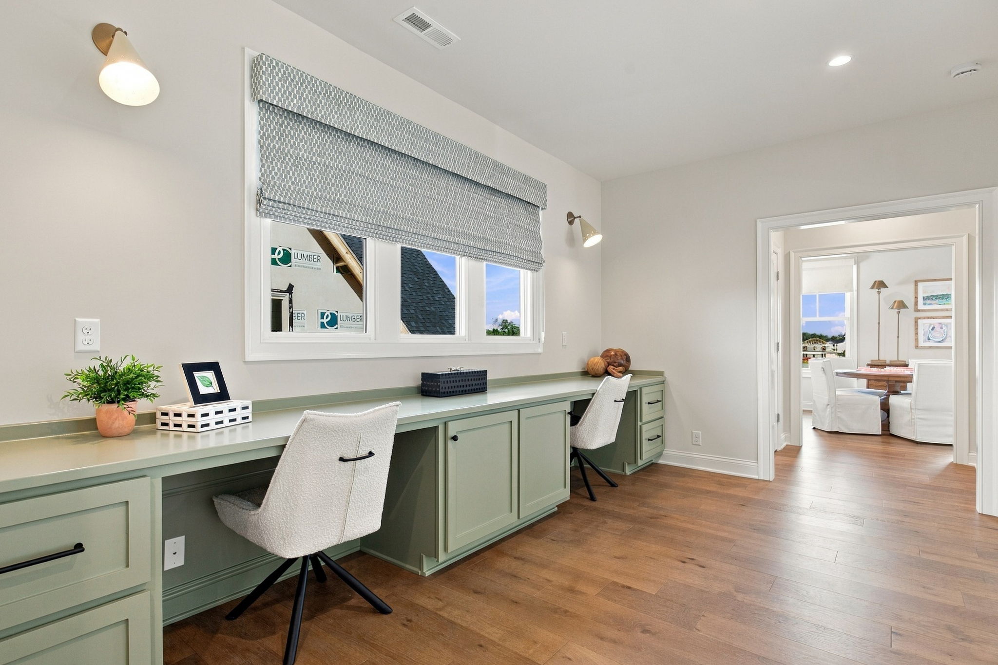 Long mint-green built-in desk with matching cabinets runs along a sunlit wall, two white swivel chairs, a potted plant, and decorative items under a patterned window shade in a bright home office.