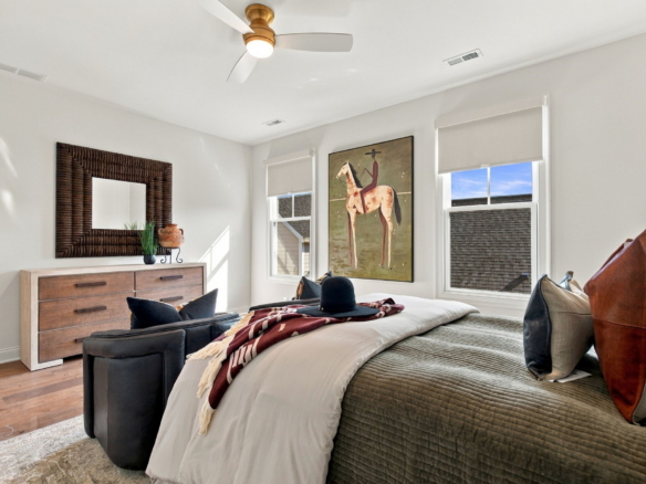 Bedroom with two twin beds, gray quilts, brown pillows, a wooden dresser, and a large horse painting between two windows under a ceiling fan.