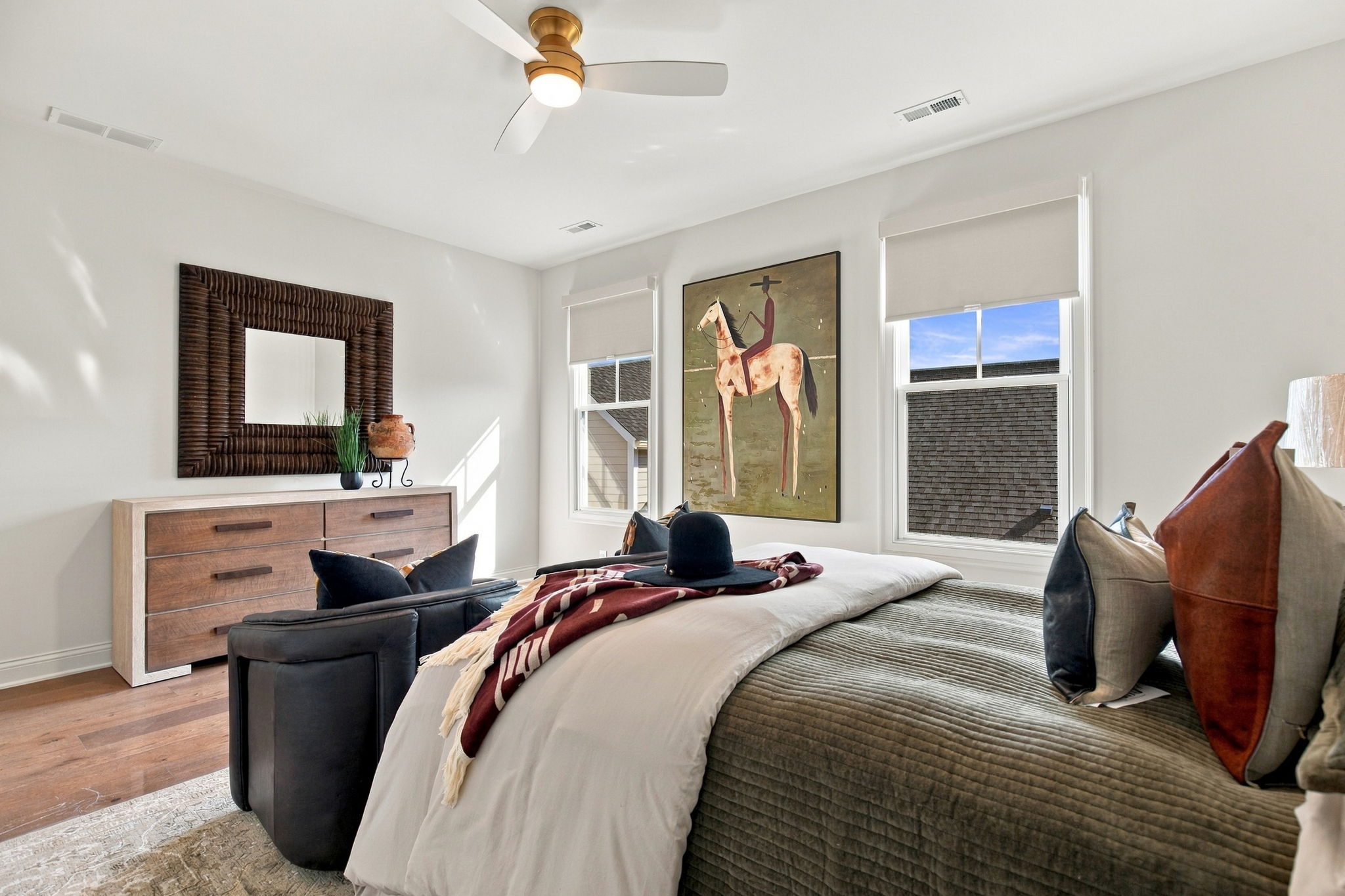 Bedroom with two twin beds, gray quilts, brown pillows, a wooden dresser, and a large horse painting between two windows under a ceiling fan.