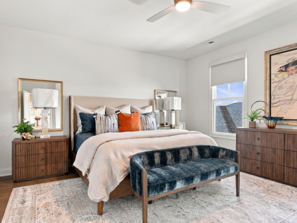 Neutral-toned bedroom with an upholstered headboard, striped and orange cushions, two wooden nightstands with lamps, large framed artwork, a window, and a blue velvet bench at the foot.