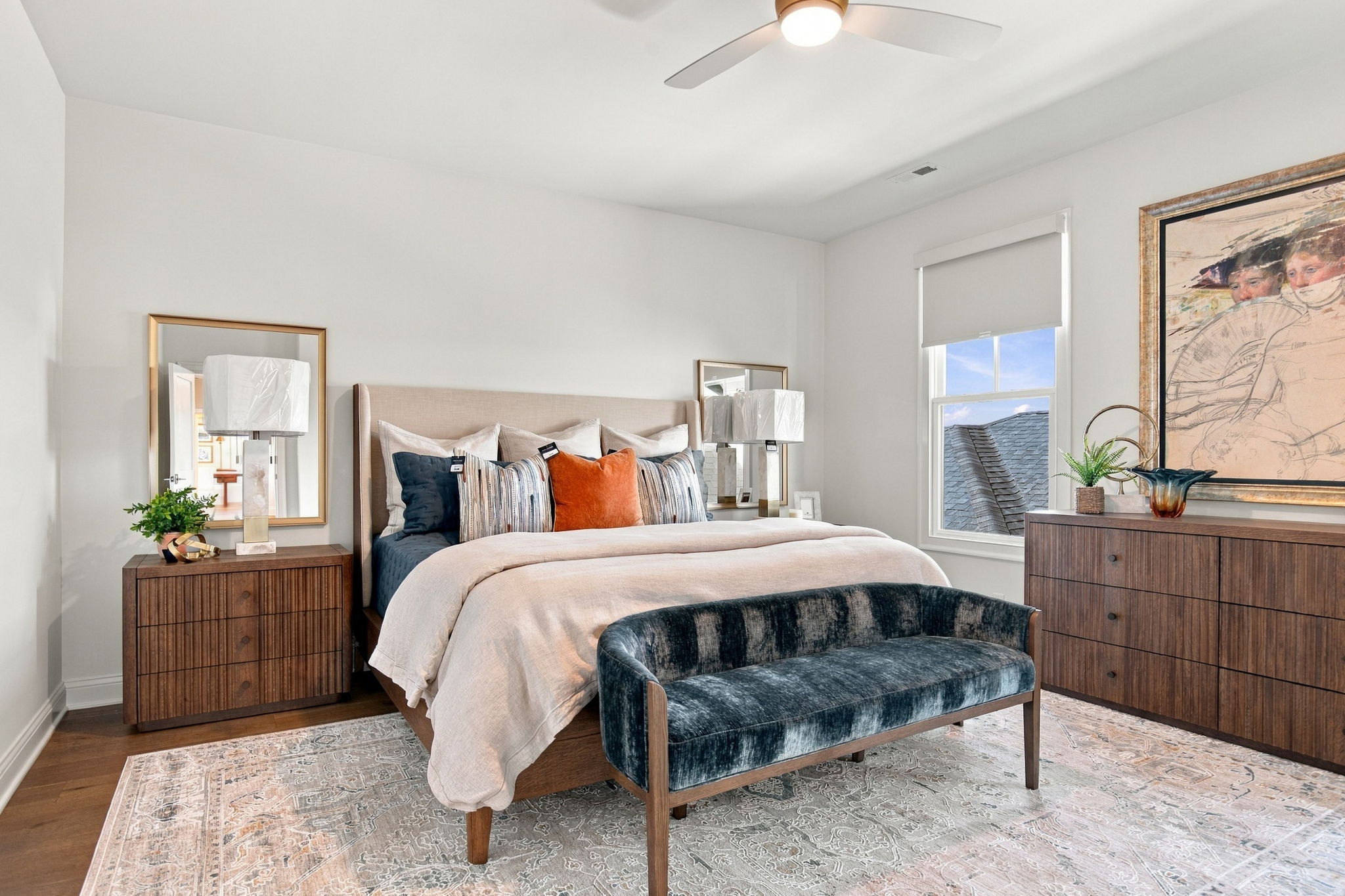 Neutral-toned bedroom with an upholstered headboard, striped and orange cushions, two wooden nightstands with lamps, large framed artwork, a window, and a blue velvet bench at the foot.