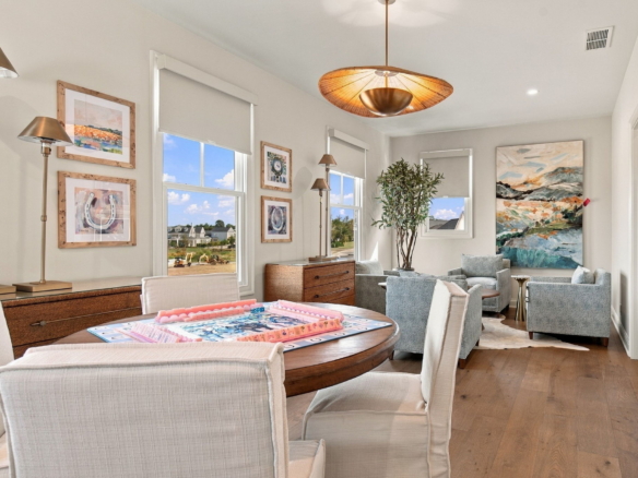 Living room with light furniture, a round wooden table set for a board game, and art on the walls by large windows.