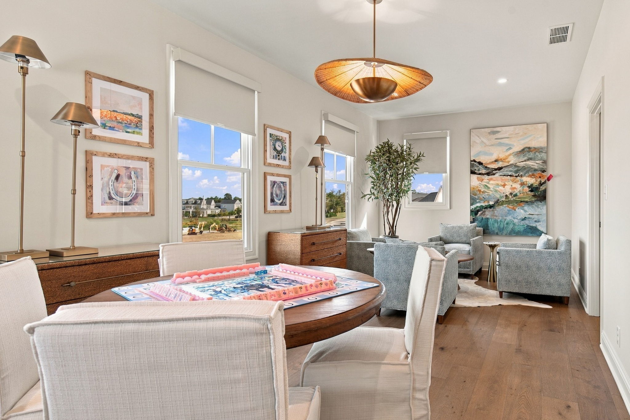 Living room with light furniture, a round wooden table set for a board game, and art on the walls by large windows.