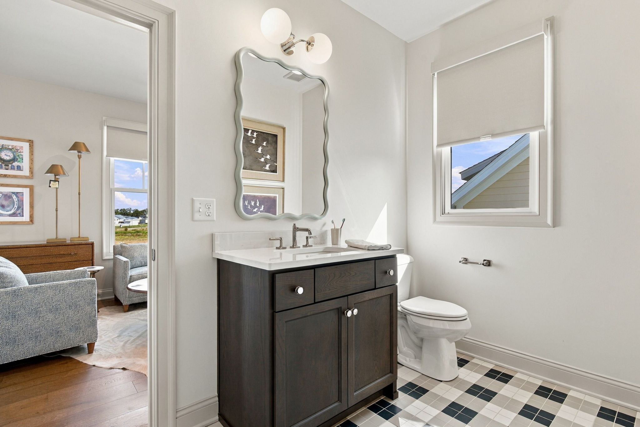 Bathroom with dark wood vanity, white countertop, wavy-edge mirror, and a two-light wall sconce above; window with blind to the right.